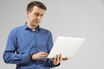 young man looks at laptop screen in his hands and stands isolated on light background