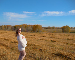 Naklejka na ściany i meble Beautiful pregnant woman in a white woolen sweater. Happy pregnant woman posing on a mown wheat field.
