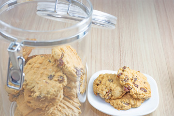 Hearty raisin cookies put on a white plate. Some of the cookies in the cookie jar lid off. The background is tiled flooring mimics wood.
