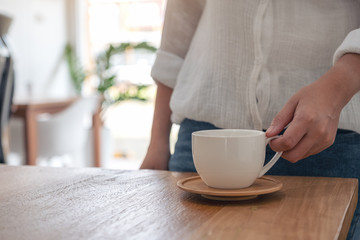 Closeup image of a woman holding a white cup of hot coffee on wooden table in cafe