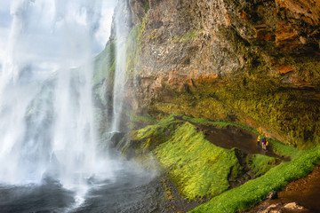 Travel in Iceland and sightseeing of waterfalls. Group of four people in colorful raincoats are standing on the trail under the rock and looking at the famous Seljalandsfoss waterfall at sunset