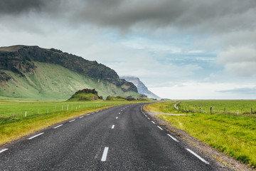 Naklejka premium Asphalt road in Iceland countryside among green fields and rocky mountains on cloudy summer day. Travel on Ring Road number 1
