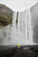 A little girl in a yellow raincoat stands in front of a huge waterfall. Iceland.
