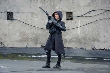 Young woman in modern black techwear style with rifle posing on the rooftop, portrait of redhead...