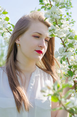 Portrait of a young beautiful woman in white top standing among blooming apple trees on a sunny day.