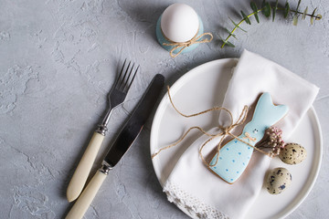 Easter  festive table setting with Easter bunny cookies, quail eggs  and chicken egg, green  leaf sprigs of eucalyptus. On a gray concrete background. Holiday celebration decorations.