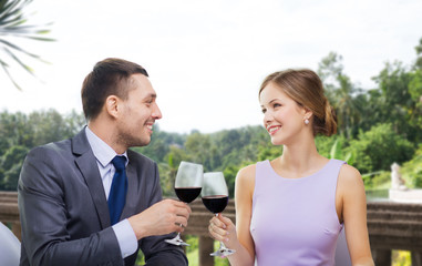 dating, celebration and valentines day concept - smiling young couple clinking glasses of non-alcoholic red wine and looking at each other at restaurant over summer background