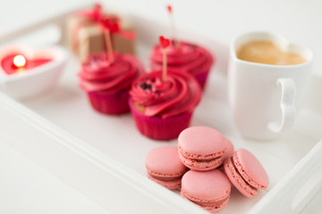 valentines day and sweets concept - close up of pink macarons, frosted cupcakes with red heart shaped cocktail sticks and coffee cup on tray