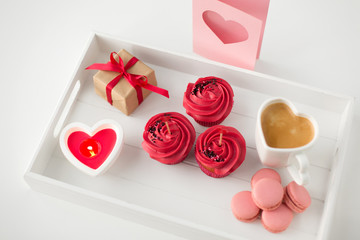 valentines day and sweets concept - close up of cupcakes with red buttercream frosting and heart shaped cocktail sticks, macarons, candle, coffee cup and gift box on tray