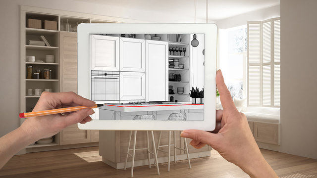 Hands holding and drawing on tablet showing modern white and wooden kitchen CAD sketch. Real finished interior in the background, architecture design presentation
