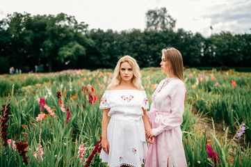 Two pretty young female friends in ethnic dresses relaxing in flower garden in summer. Couple of cute barefoot women in country embroidery holding hands and hugging outdoor. Stylish fashionable girl