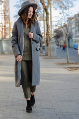 Outdoor portrait of young beautiful fashionable woman posing in street. Model wearing stylish gray coat, women's suit with trousers, Fedora hat. Female fashion concept, city lifestyle