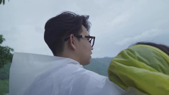 Slow motion - Young Asian sweet couple feeling happy using romantic time playing rain while wearing raincoat standing near lake. Lifestyle couple enjoy and relax in rainy day.