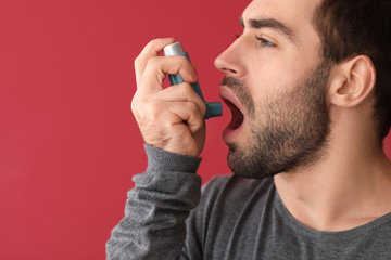 Young man with inhaler having asthma attack on color background