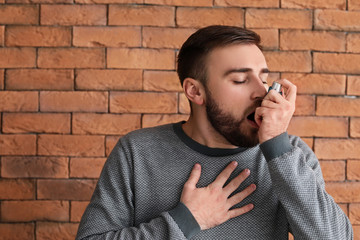Young man with inhaler having asthma attack against brick wall