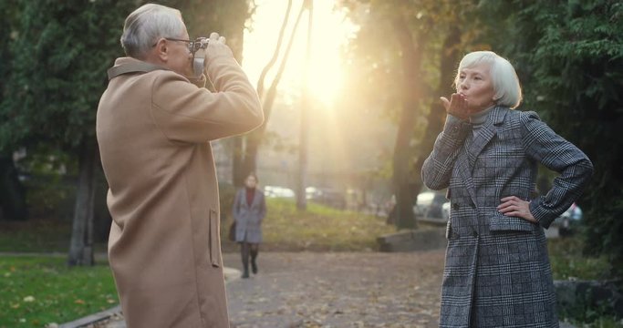 Old Caucasian man taking photo on the camera of his lovely grey haired wifi in the park in autumn. Outside.