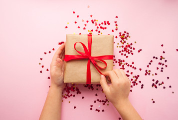 Child is holding gift box wrapped in brown colored craft paper and tied with red bow on pink background with red heart shape confetti.