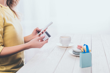 cropped view of woman holding smartphone and credit card near toy shopping bag on table