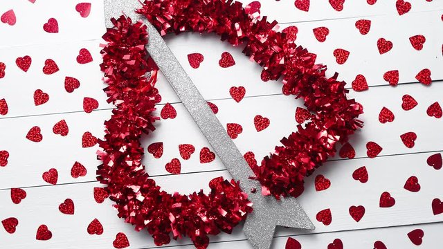 Valentine's Day decoration composition. Heart with arrow as a love symbol placed on background with red sequins on white wooden table. Romantic background. Flat lay, top view.