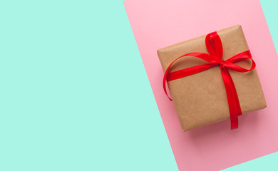 Gift box wrapped in brown colored craft paper and tied with red bow on pink and turquoise background with copyspace.