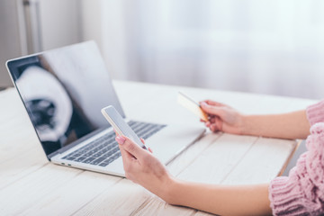 cropped ciew of woman holding credit card and smartphone near laptop