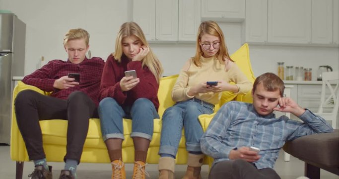 Indifferent teenage friends spending leisure in domestic room, obsessed addicted with smart phones ignoring each other. Attractive teenagers phubbing each other, networking online on cellphone at home