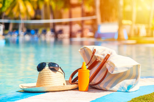 Straw hat with sunglasses near sunscreen lotion bottle and beach bag against the surface of blue clear swimming pool. Summer vacation concept.