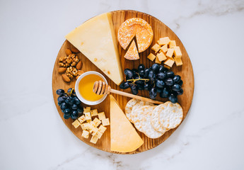 Flat lay with various types of cheese, grapes, nuts, honey and cracker in wooden board on marble
