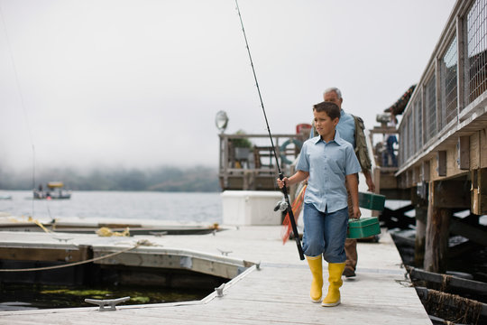 Boy walking along a jetty carrying fishing equipment with his father.