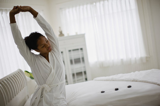 Young woman sitting on her bed and stretching while wearing a white robe.