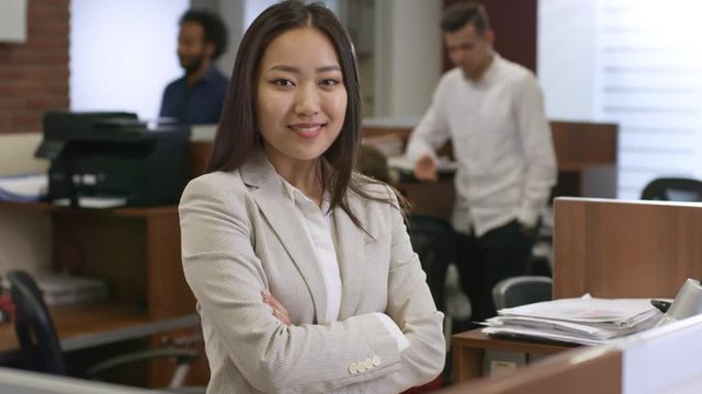 Portrait of cheerful young Asian woman in formal clothes standing with arms crossed and smiling at camera while colleagues working in the background, medium shot