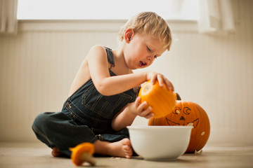 Young boy sitting cross-legged on a floor pours seeds out of a small pumpkin into a bowl with a big pumpkin with a face drawn on it in the background.
