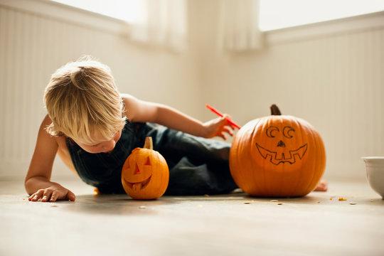 Young boy lies on his side and looks down at a small Jack O'Lantern next to a big,  uncarved pumpkin with a face drawn on it.