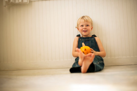 Young boy holds a small Jack O'Lantern on his knees as he sits on a floor leaning against a wall and poses for a portrait.