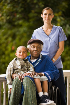 Senior man in a wheelchair with his grandson sitting on his lap and a female nurse standing behind them pose for a portrait on a porch.