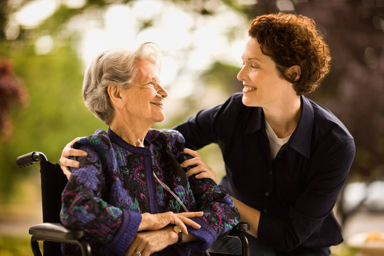 Mature woman leans over to smile and talk to a senior woman with a oxygen nose hose (cannula) sitting in a wheelchair outside.