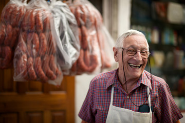 Smiling senior man stands in his butcher's shop with sausages hanging in the background.