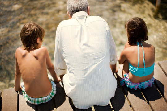 Old grandfather sitting with his two young grandchildren on a wooden jetty at the beach.