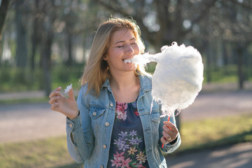 charming caucasian girl walks in the spring Park in a denim jacket and eats pink cotton candy,...