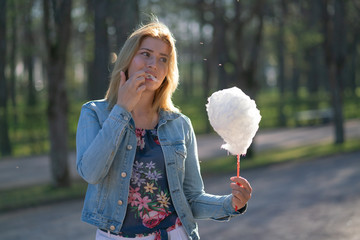 charming caucasian girl walks in the spring Park in a denim jacket and eats pink cotton candy,...