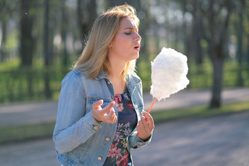 charming caucasian girl walks in the spring Park in a denim jacket and eats pink cotton candy,...