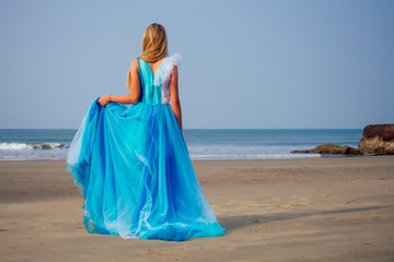 Fototapeta na wymiar Young beautiful caucasian blonde woman in long tull butterfly white blue dress on coast of sea.female running towards the ocean freedom