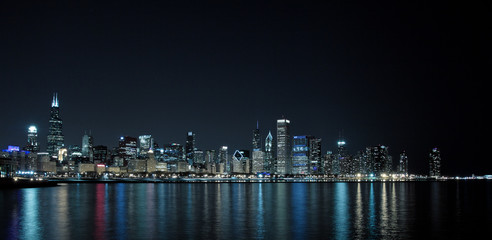 Long-exposure photo of downtown Chicago at night. The light of the stars can be see in the clear sky above, and the reflection of the city lights is in the lake in the foreground.