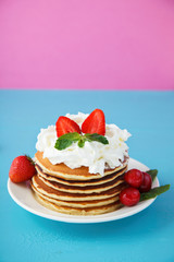 pancakes in a plate with whipped cream, honey, strawberries, mint and sweet cherries on a bright blue and pink background.