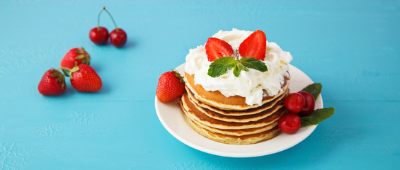 pancakes in a plate with strawberries, mint, honey and cherry on a bright blue background.