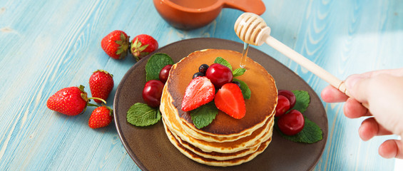 pancakes in a plate with strawberries, mint, honey and cherry on a bright blue background.
