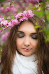 Fototapeta premium Beautiful blonde girl standing in a white scarf and closed her eyes in the garden of cherry blossoms. Vertical photography