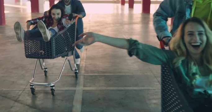 Joyful young Caucasian hipsters having fun. Two attractive joyful girls sitting in the carriages while being carried by their boyfriends in the empty parking. Indoors