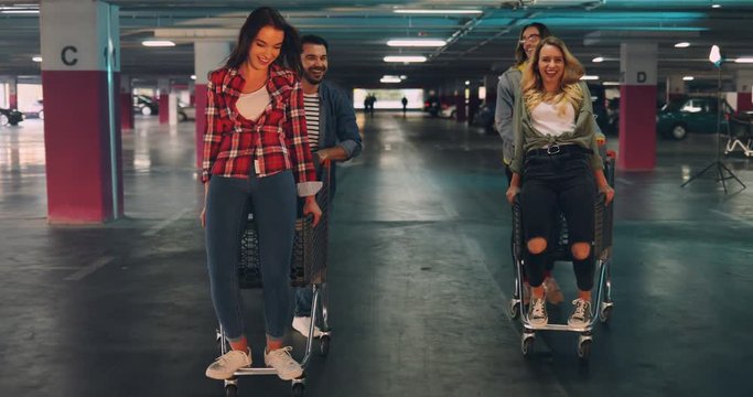 Two cheerful caucasian couples of young hooligans having fun at the parking space underground while riding the trolleys. Indoors.