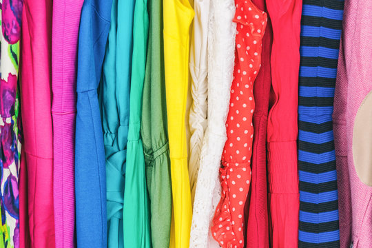 Closet women's fashion outfits clothes arranged in rainbow colors assorted. Clothing store dresses hanging on shopping rack. Variety of fabrics and patterns, wool, polyester, polka dots, stripes.